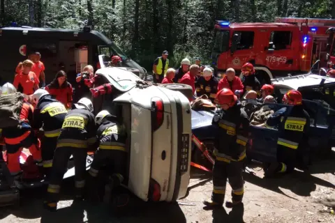 Akcja ratunkowa po wypadku. Strażacy KSRG zabezpieczają teren i wyciągają poszkodowanych z przewróconego auta. W tle wozy strażackie i karetka.