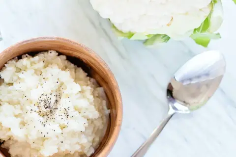 Pyszne puree z kalafiora w drewnianej misce, posypane pieprzem. Obok leży łyżka, a w tle świeży kalafior.