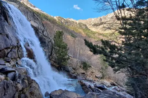 La cascade des Molières dévale une paroi rocheuse, entourée de pins et de montagnes enneigées sous un ciel bleu.