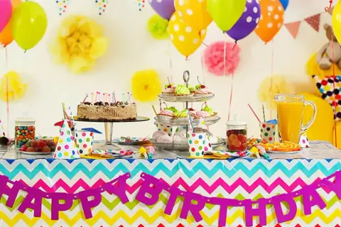 Kolorowy stół z tortem, babeczkami i słodyczami czeka na imieniny Gosi. Balony i girlanda "Happy Birthday" tworzą radosną atmosferę.