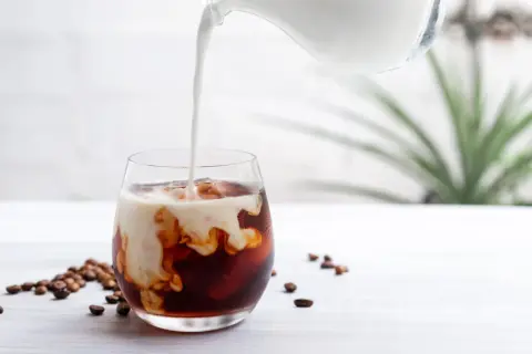 Mjölk hälls i ett glas med iskaffe, perfekt för ett enkelt iskaffe recept. Kaffebönor ligger utspridda bredvid.