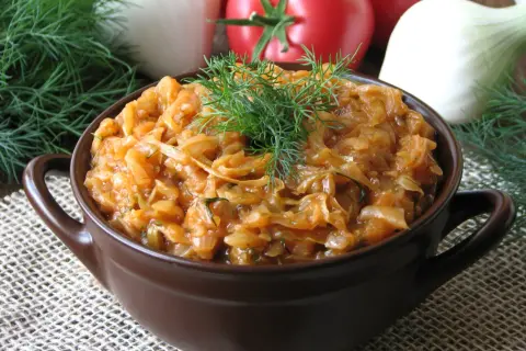Młoda kapusta z pomidorami, duszona z koperkiem, podana w brązowej miseczce. Na tle widać pomidory i cebulę.