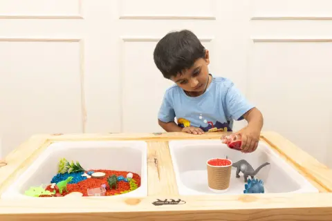 A boy pours red liquid into a sensory table with dinosaurs and colorful rice, exploring fun sensory table ideas.