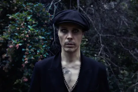 Ville Valo w czarnym kaszkiecie i marynarce, z intensywnym makijażem oczu, stoi na tle ciemnej, leśnej roślinności.