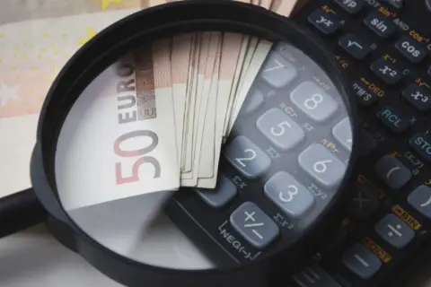 Lupe über 50-Euro-Scheinen und einem Taschenrechner. So wird die Frage "wie viel verdient man im Referendariat" visuell beleuchtet.