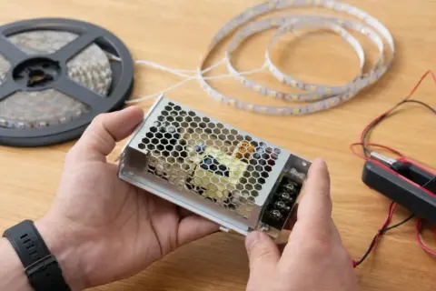 Ręce trzymają zasilacz do taśmy LED. W tle szpulka z taśmą i miernik. Nowoczesna technologia LED gotowa do montażu.