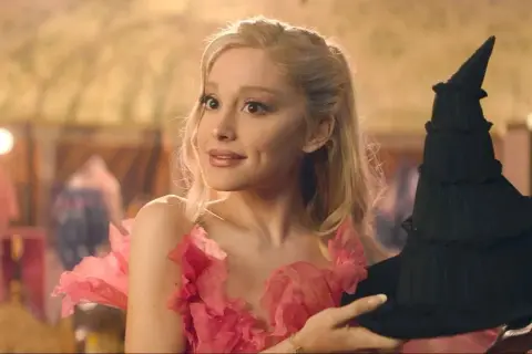 Ariana Grande w różowej sukni z kwiatami trzyma czarny kapelusz. Scena z jej filmów.