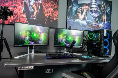 Ein Streamer-Setup: Zwei Monitore, Mikrofon, Kamera auf Stativ, Gaming-PC mit blauer Beleuchtung und Bilder von E-Sport-Erfolgen. So macht ein Streamer seine Inhalte.