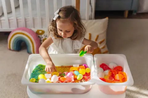 A young girl explores colorful rice and toys in sensory bins, demonstrating why sensory bins are important for early childhood development and learning through play.