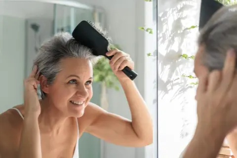 Mujer sonriente se peina el cabello gris en el espejo, lista para matizar canas y lucir radiante.