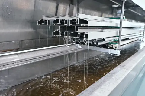 Profili in alluminio immersi in un bagno per la sverniciatura chimica, gocciolando liquido marrone.