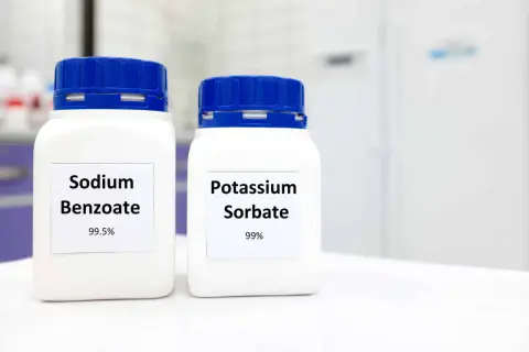 Zwei weiße Behälter mit blauen Deckeln stehen nebeneinander. Auf dem linken Behälter steht "Sodium Benzoate 99.5%", auf dem rechten "Potassium Sorbate 99%".