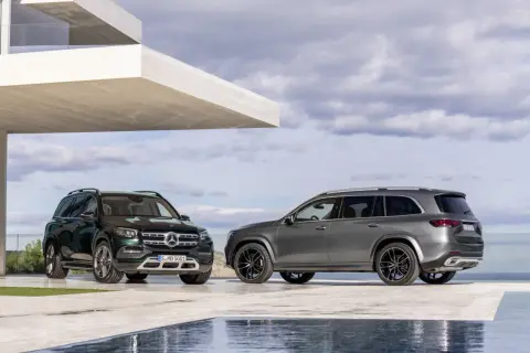 Dwa Mercedesy GLS 2025, jeden zielony, drugi szary, zaparkowane przed nowoczesnym budynkiem z basenem.
