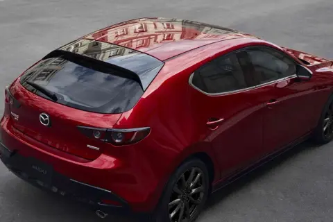 Czerwony hatchback Mazda 3 z czarnymi felgami, odbijający budynek na dachu. Wygląda elegancko, w przeciwieństwie do nudnego sedana.