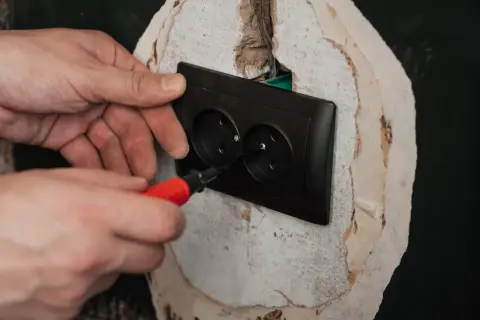 Ręce montują czarne podwójne gniazdko elektryczne. Śrubokręt z czerwoną rączką dokręca śrubę, kończąc podłączenie gniazdka.
