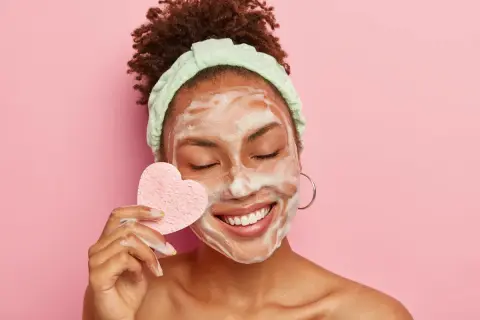 Mujer sonríe con espuma en la cara, usando una esponja en forma de corazón para su limpieza facial.