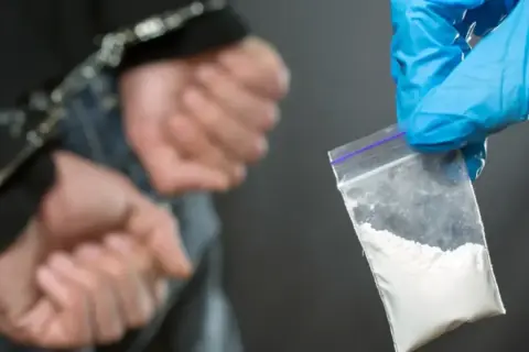 Ręce w kajdankach, obok woreczek z białym proszkiem, prawdopodobnie klefedron.