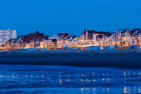 Kvällsljus över strandpromenaden i Dunkirk stad. Husen längs kusten lyser upp mot den mörka himlen, med reflektioner i det lugna vattnet.