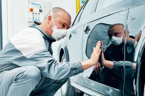 Un operaio con mascherina controlla la carrozzeria lucida di un'auto, pronto per carteggiare auto e rifinire la verniciatura.