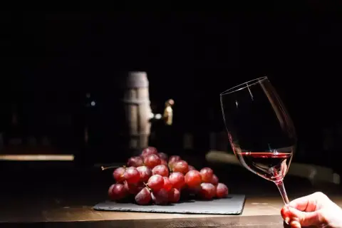Czerwone winogrona i kieliszek wina, a w tle beczka. Czy wino bezalkoholowe ma alkohol? To pytanie nurtuje wielu miłośników tego trunku.