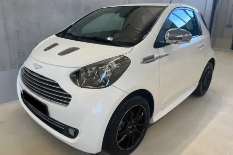Biały Aston Martin Cygnet z czarnymi felgami i chromowanymi detalami, prezentujący się elegancko w garażu.