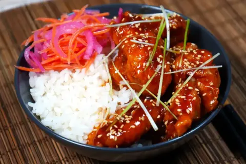 En skål med ris, krispigt koreanskt friterad kyckling i en söt och kryddig sås, toppad med sesamfrön och salladslök. Serveras med en sida av picklad morot och rödlök.