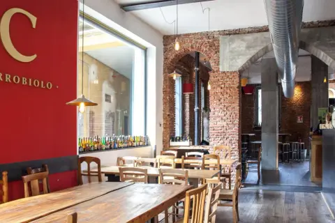 Interno di un locale con tavoli in legno e mattoni a vista. Sulla parete rossa, il logo "C CARROBIOLO" e una vetrina con tante bottiglie di birra.