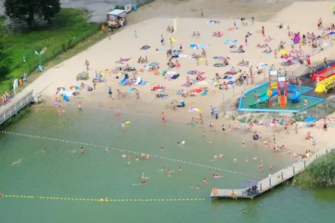 Wakacyjny dzień nad jeziorem margonińskim. Ludzie opalają się na plaży, kąpią w wodzie i korzystają ze zjeżdżalni wodnych.