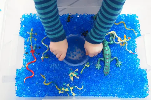 Kinderhände erkunden eine Kiste voller blauer Wasserperlen mit Spielzeug-Echsen und Schlangen. Tolle Wasserperlen Ideen für sensorisches Spiel!