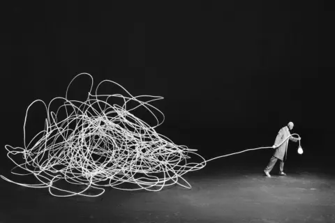 Człowiek z żarówką, jak w pracach Gilbert Garcin, ciągnie za sobą plątaninę białego sznurka na czarnym tle.