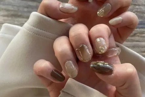 Manucure coréenne aux tons bruns et beiges, avec des accents dorés et des bagues transparentes.