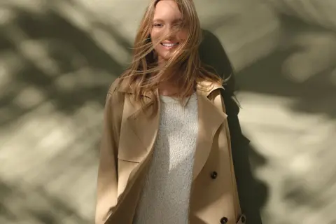 Uśmiechnięta blondynka w beżowym trenczu i swetrze, z rozwianymi włosami. Inspiracja, jak nosić trencz w wiosenny dzień.