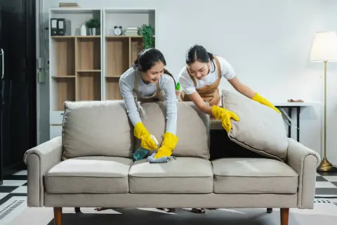 Dos mujeres con guantes amarillos limpian un sofá beige. Una rocía limpiador y frota con un paño, la otra levanta un cojín.