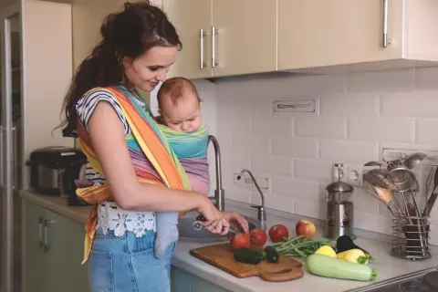 Mamma förbereder mat vid amning i köket, med en bebis i bärsjal. Grönsaker ligger på bänken.