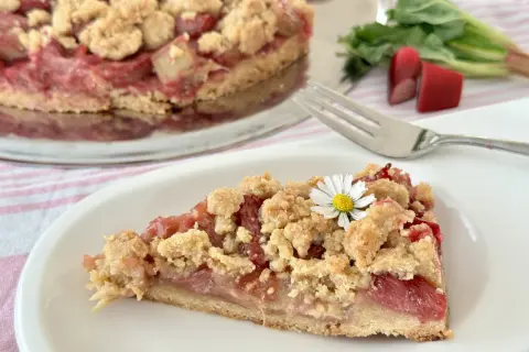 Ein saftiges Stück Rhabarberkuchen mit Schmand und Streuseln, garniert mit einer kleinen Gänseblümchen.
