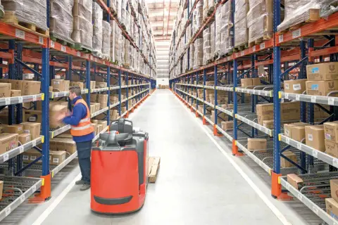 Pracownik magazynu, order picker, przenosi paczkę. Czy ciężko? Wysokie regały pełne towarów.