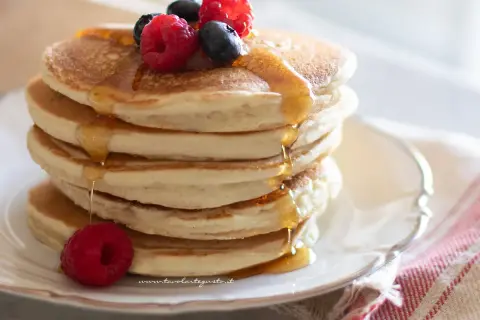 Pancakes senza uova dorati, impilati e inzuppati di sciroppo, guarniti con lamponi e mirtilli freschi.