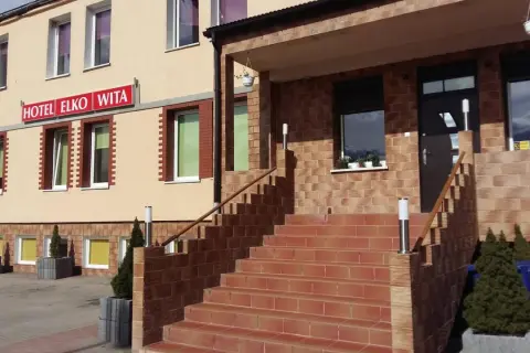 Wejście do hotelu Elko Elbląg. Schody prowadzą do drzwi, a nad nimi widnieje czerwony szyld.