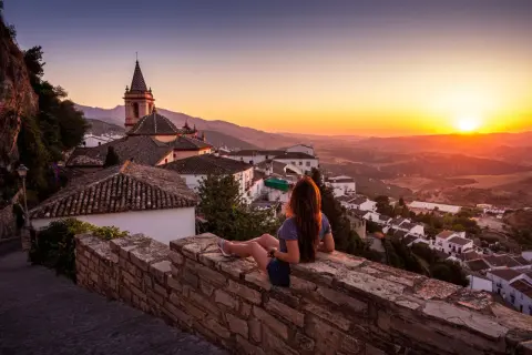 Una mujer contempla el atardecer sobre un pueblo blanco, con tejados de los mayses y montañas al fondo.