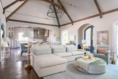 A spacious living room with a sectional sofa, armchair, and ottoman. The vaulted ceiling with exposed beams and a large chandelier create a dramatic focal point, showcasing what is the best way to arrange furniture for an open, airy feel.