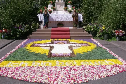 Blumenbild mit Kreuz und Schriftzug "Ich bin das Licht der Welt" vor einem Altar. Ein Fest für Fronleichnam.