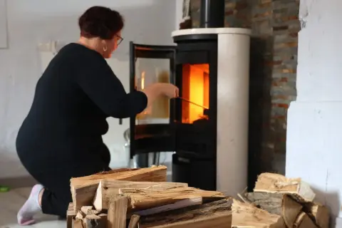 Frau kniet vor einem brennenden Kamin, um ihn zu reinigen. Holzscheite liegen bereit.