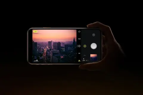 Dłoń trzyma smartfon z widokiem na miasto o zachodzie słońca. Na ekranie widać interfejs aparatu, gdzie można ustawić ISO.