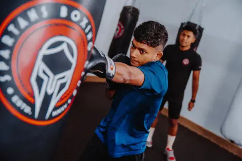 A young boxer practices his boxing combos, throwing a punch at a heavy bag in a gym. Another person watches in the background.
