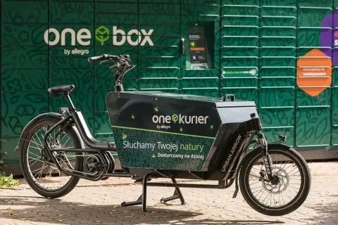 Rower cargo z napisem "one kurier by allegro" stoi przed zielonym paczkomatem "one box".