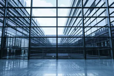Interior de un edificio moderno con amplios muros cortina de cristal que reflejan el cielo y la arquitectura circundante.