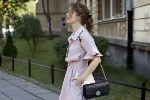 Kobieta w różowej sukience i czarnej torebce na łańcuszku na tle budynku i zieleni.