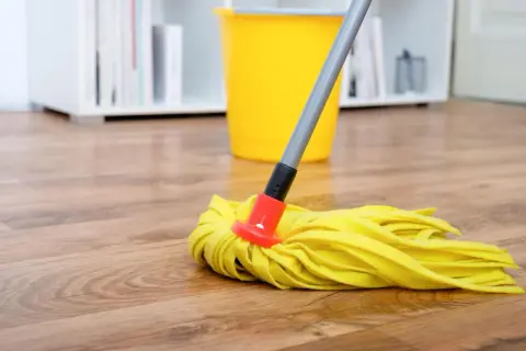 Ein gelber Wischmopp liegt auf einem Holzfußboden, bereit, den Boden zu wischen. Ein gelber Eimer steht im Hintergrund.