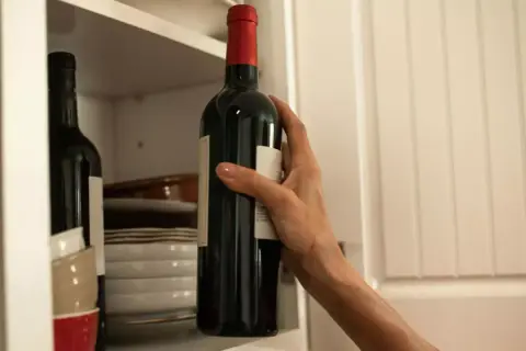Dłoń sięga po butelkę bordeaux wina z półki, obok stosu talerzy i miseczek.