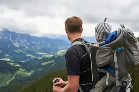 Mann mit Kind auf dem Rücken in einer Kraxe, blickt auf bergige Landschaft.